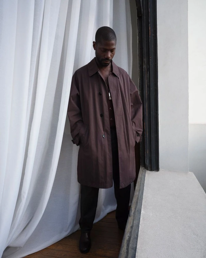 Model in a long brown coat standing next to white curtains near a doorway for Lemaire lookbook