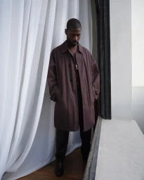 Model in a long brown coat standing next to white curtains near a doorway for Lemaire lookbook