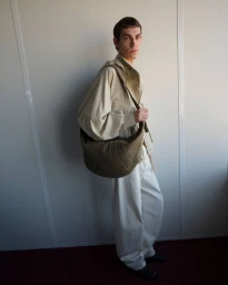 Model wearing a light utility shirt and white trousers carrying a large curved canvas shoulder bag against a neutral studio wall.