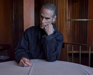 Man wearing a dark jacket seated at a table with a white tablecloth against wood paneling