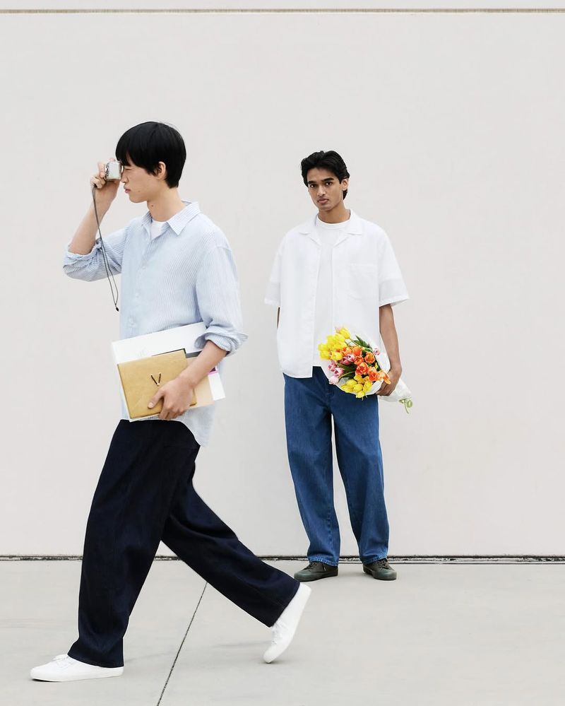 Two young men pose outdoors; one holds flowers and the other holds a camera. One man walks toward the camera in a relaxed pose.