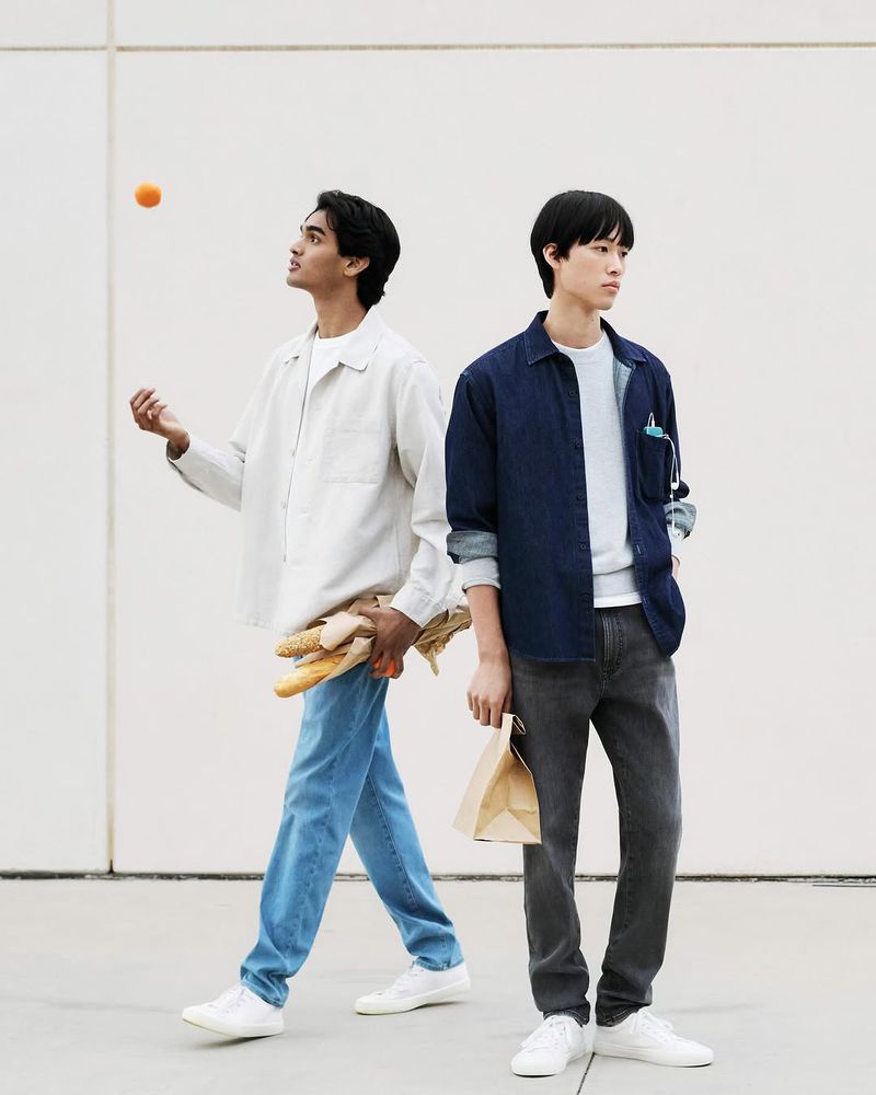 Two young men pose outdoors; one juggles an orange and holds bread, while the other carries a paper bag. They wear casual outfits.