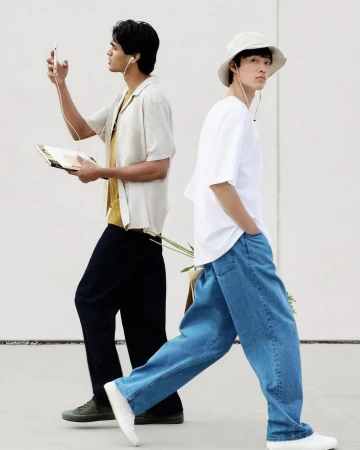 Two young adult men in casual fashion pose back to back against a light gray wall, one holding a phone and the other in wide-leg jeans.
