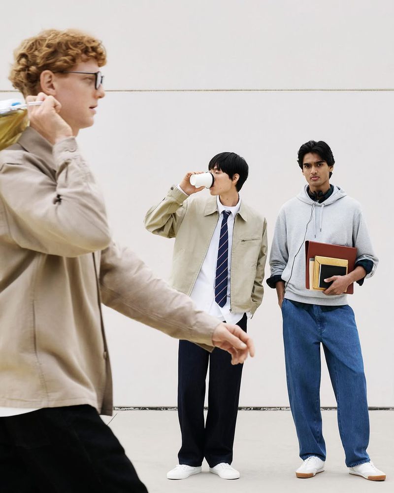 Three young adult men standing in front of a neutral wall, one drinking from a paper cup, another carrying books.