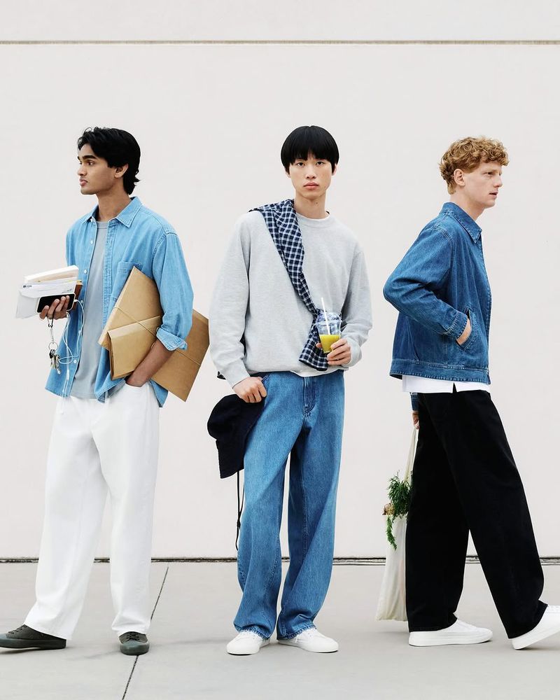Three stylish men in denim and casual wear pose against a white wall, showcasing contemporary fashion and diverse looks in an urban setting.