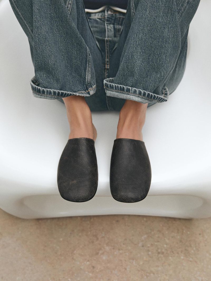 Person sitting on a white chair wearing blue jeans and black leather mules in an eye-level shot indoors.