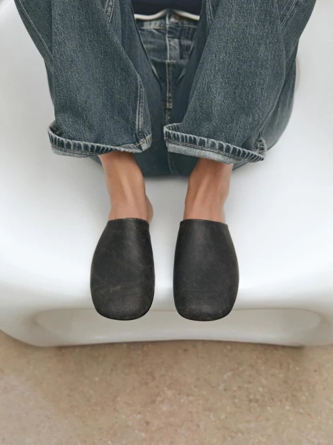 Person sitting on a white chair wearing blue jeans and black leather mules in an eye-level shot indoors.