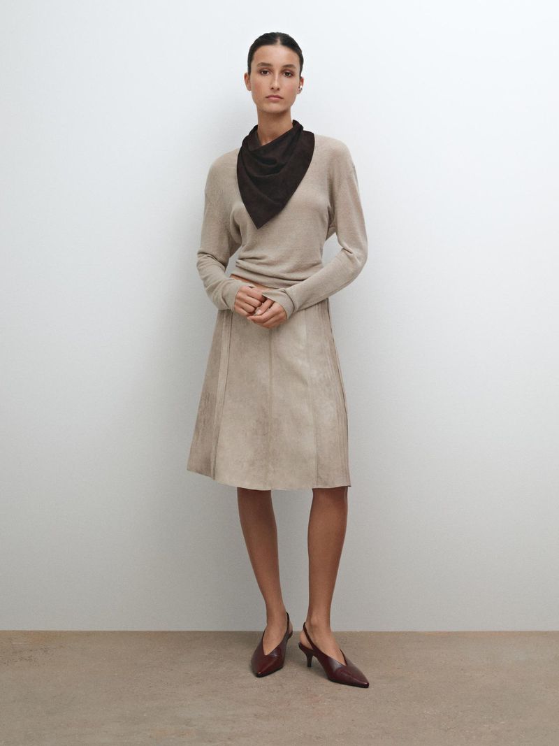 Full-body studio shot of a woman in a beige sweater, suede skirt, and burgundy kitten heels, accessorized with a dark brown neck scarf.