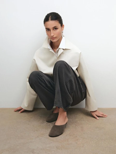 Indoor studio shot of an adult female model in a cream-colored shirt and grey pants, squatting on a neutral-tone surface.