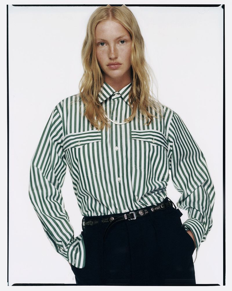 Blonde woman in a green and white striped button-down shirt and black pants posing against a white background in a fashion editorial style.