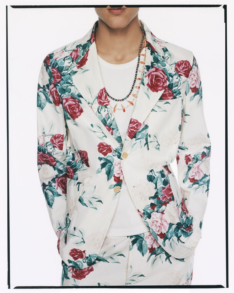 Person wearing a floral-print suit and two necklaces, posing indoors against a white background in a fashion editorial style.