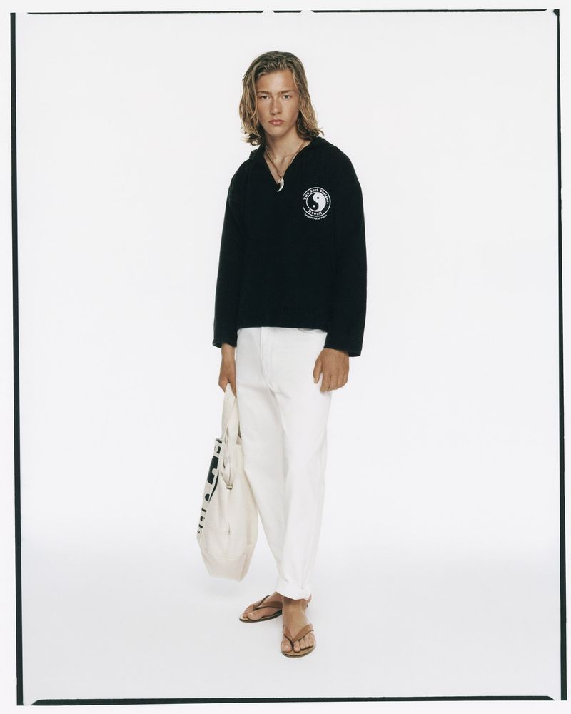 A young adult male stands in a full shot wearing a dark sweater, white pants, and sandals, holding a tote bag against a white backdrop.
