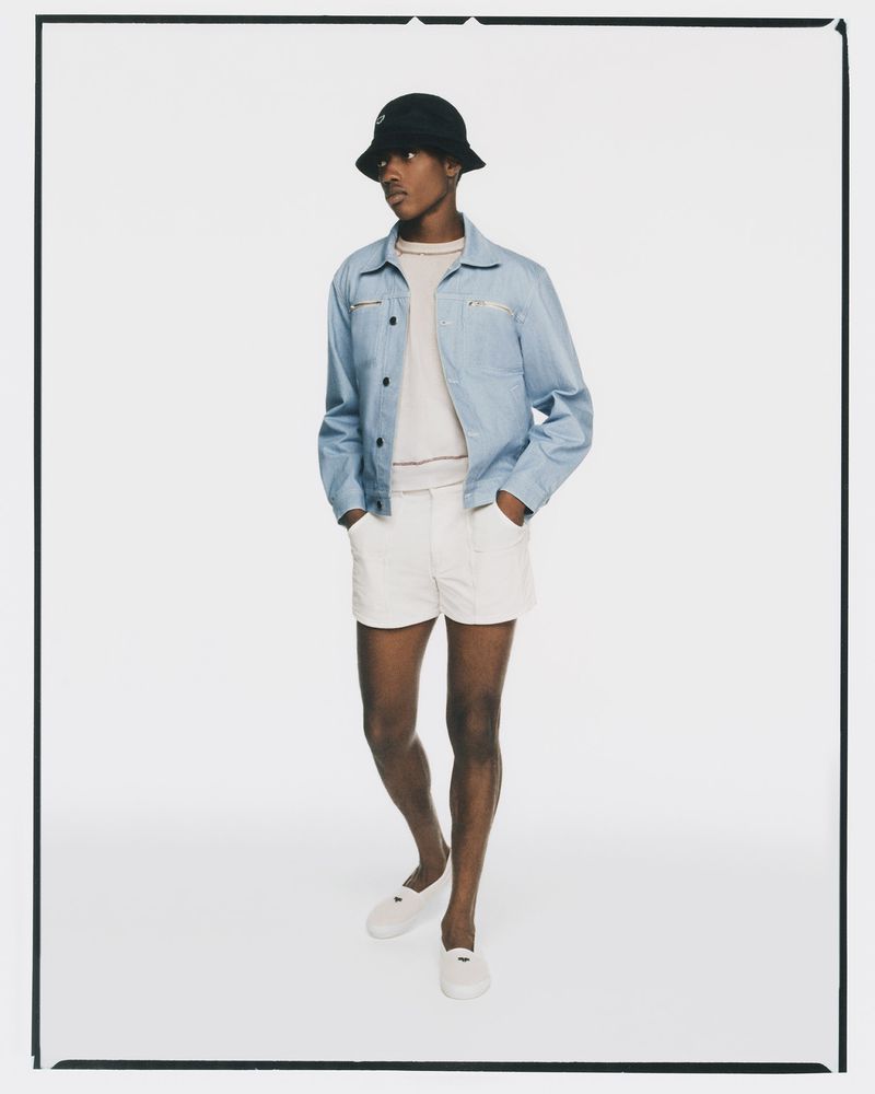 Full-length shot of an adult male in a light-blue jacket and cream shorts, wearing a dark bucket hat and slip-on shoes, standing in a white photo studio.