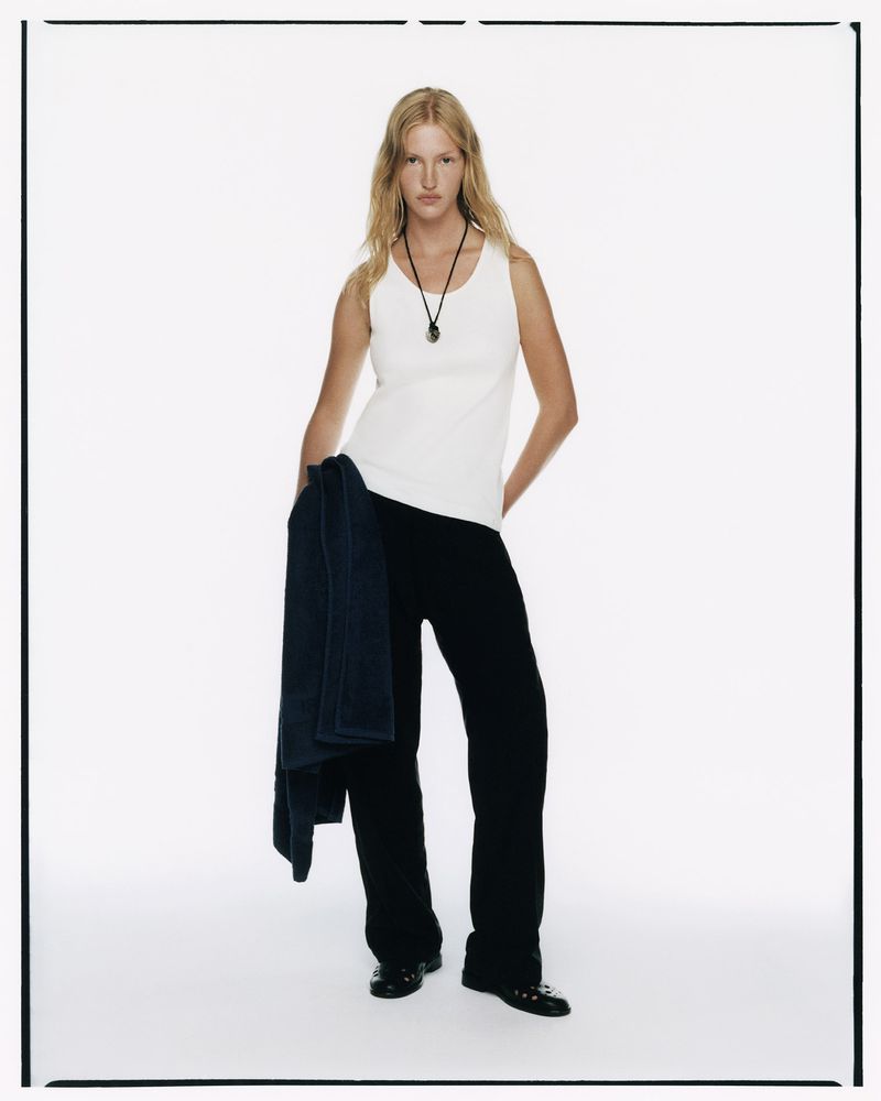 Full-length studio portrait of a female model with long blonde hair, wearing a white tank top and black pants, holding a jacket.