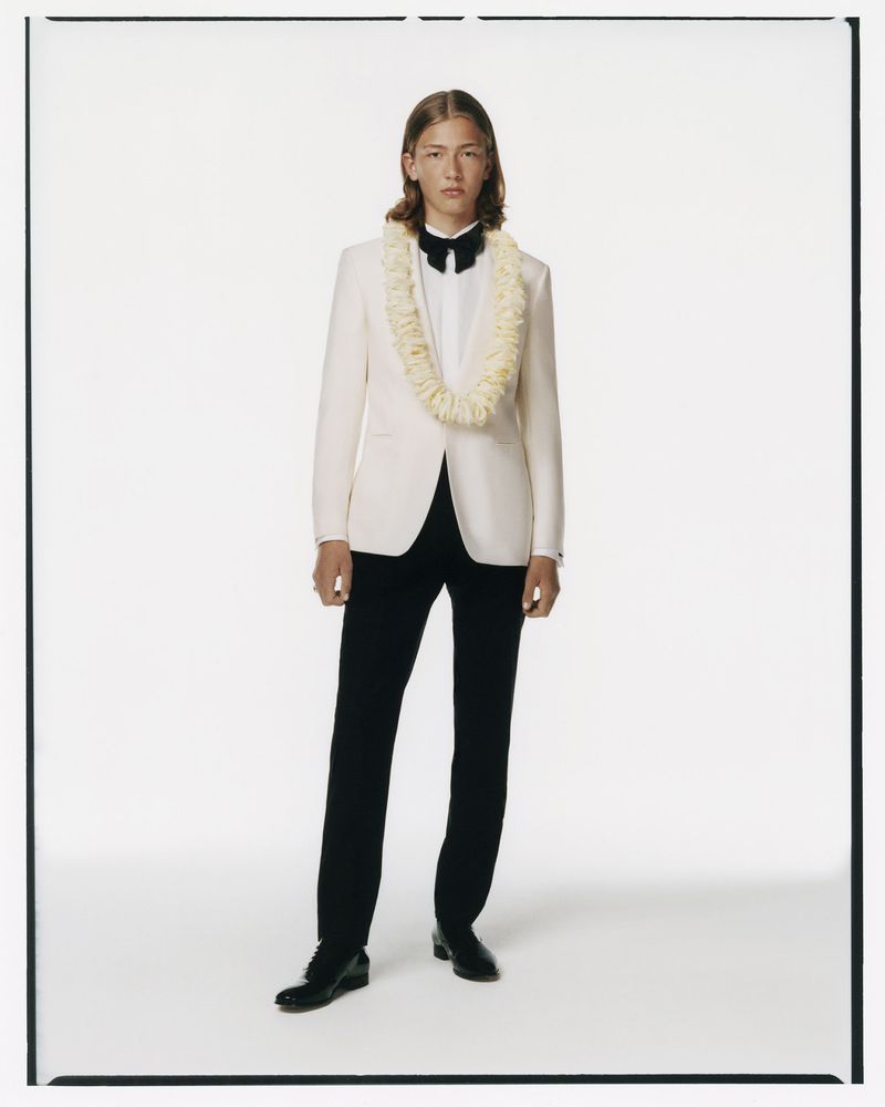 Full-length studio shot of a male model in a formal white tuxedo jacket, black pants, bow tie, and lei necklace, against white backdrop.