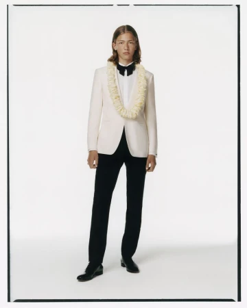Full-length studio shot of a male model in a formal white tuxedo jacket, black pants, bow tie, and lei necklace, against white backdrop.