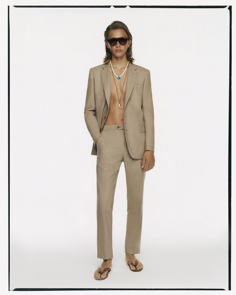 Male fashion model in light brown suit, sunglasses, and flip-flops poses in a white photo studio, showcasing summer elegance.
