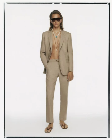 Male fashion model in light brown suit, sunglasses, and flip-flops poses in a white photo studio, showcasing summer elegance.