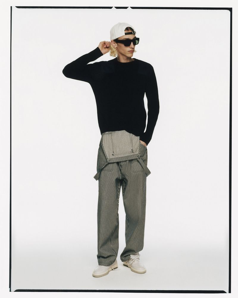 Full-length shot of a young adult male wearing a black sweater, striped overalls, white baseball cap, and black sunglasses on white.