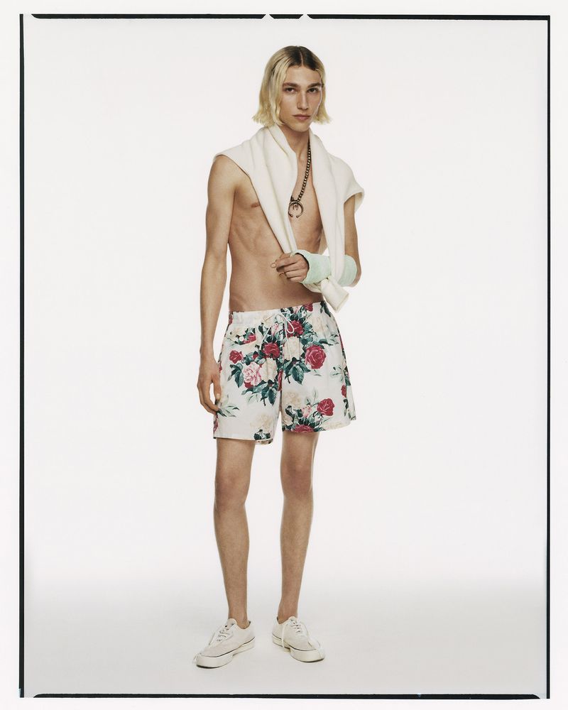 Adult male model in floral shorts and a cast on his arm, standing in a photo studio with a white background and wearing neutral-colored sneakers.