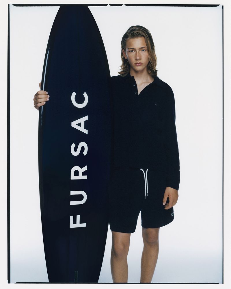 Young adult male model in black polo shirt and shorts stands holding a black surfboard with the brand name 'Fursac' visible.