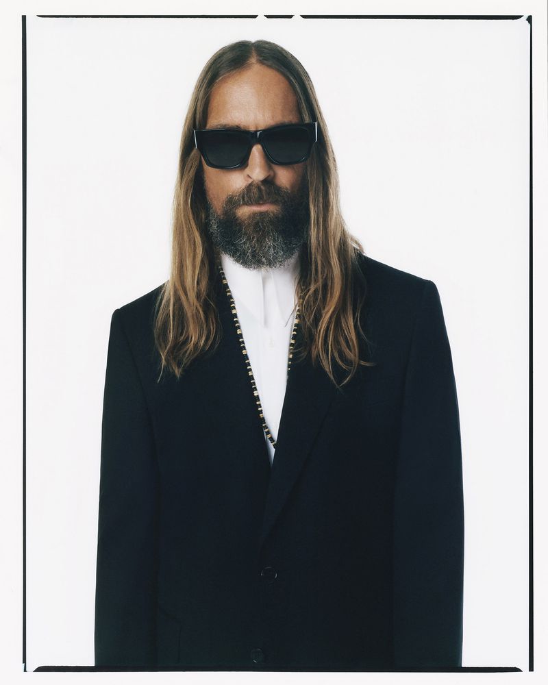 Portrait of an adult man with long hair, a beard, and sunglasses, wearing a black suit against a bright, neutral background.