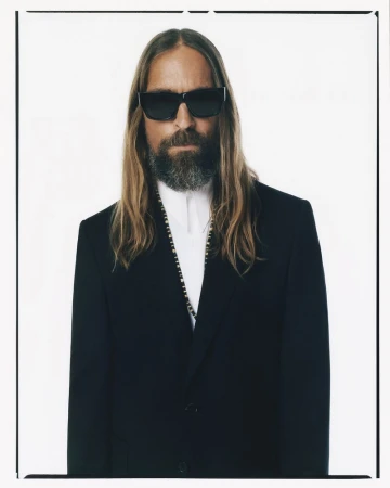 Portrait of an adult man with long hair, a beard, and sunglasses, wearing a black suit against a bright, neutral background.