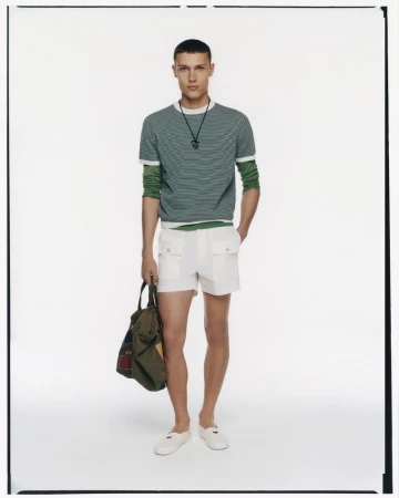 A male model poses in a studio wearing a green and white striped shirt, white shorts, and white shoes, holding an olive green tote bag.