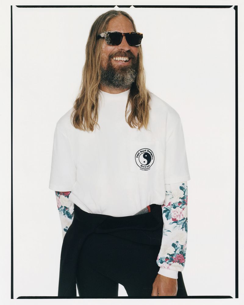Smiling man with long hair and sunglasses in white shirt with logo, floral long sleeves, dark pants, standing in a white studio.
