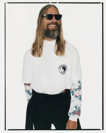 Smiling man with long hair and sunglasses in white shirt with logo, floral long sleeves, dark pants, standing in a white studio.