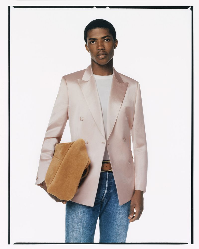 Fashion shot of a male model in a blush double-breasted blazer, white t-shirt, and jeans holding a tan suede clutch against a white backdrop.