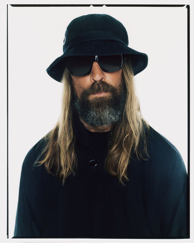 Portrait of a bearded man with long blonde hair, wearing sunglasses and a hat, against a bright white background in a studio setting.