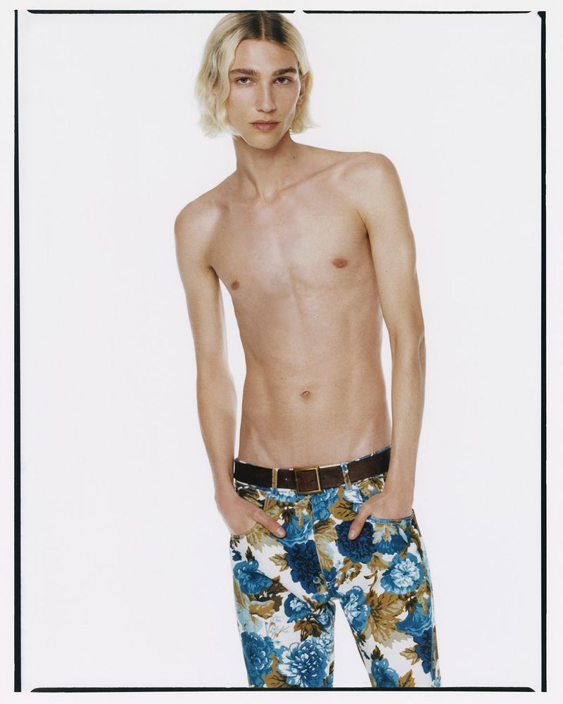A fair-skinned young adult shirtless man poses in front of a white backdrop wearing floral-patterned jeans and a brown belt. Fashion shot.