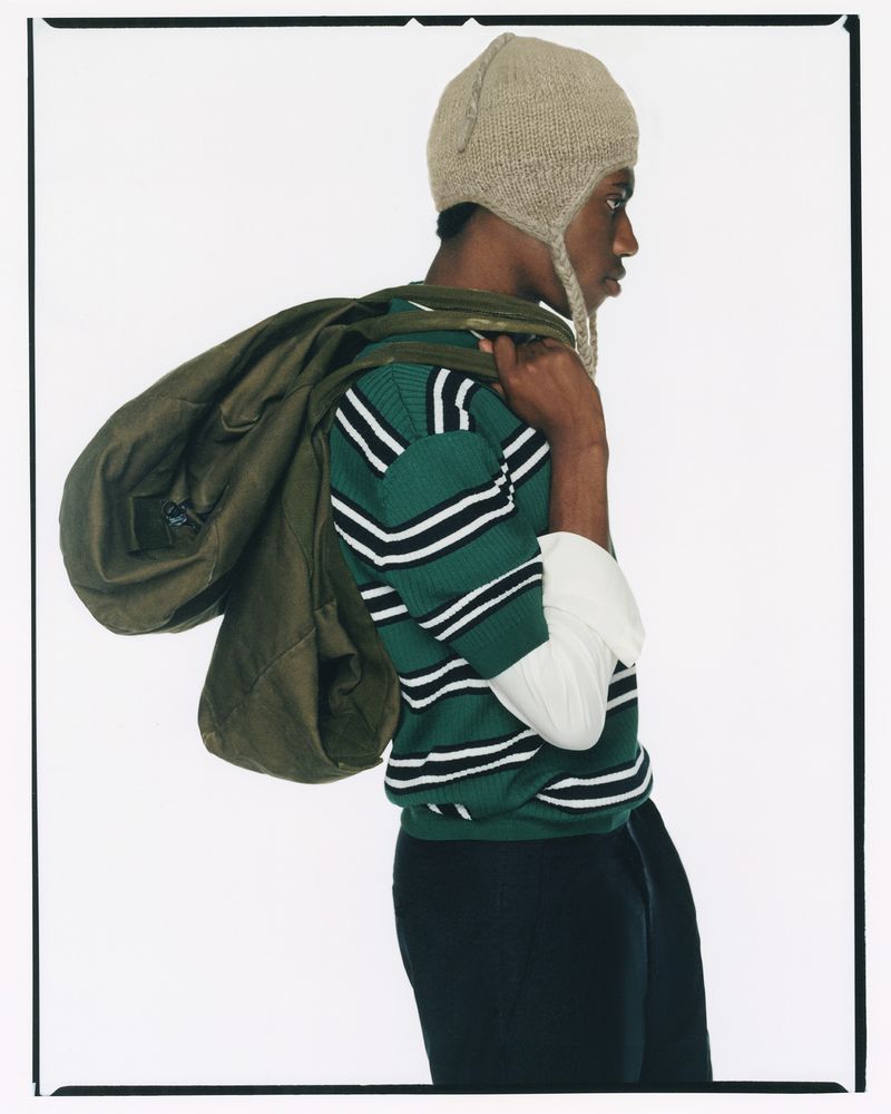 An African-American man wearing a striped green and white shirt, black pants, and a beige knit hat with a green duffel bag over his shoulder.