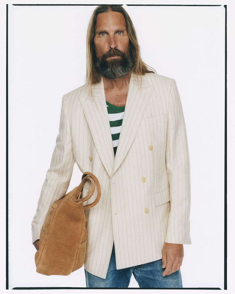 Portrait of adult male with long hair and beard wearing blazer, jeans, and carrying a brown leather tote bag on white background.