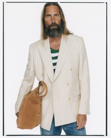 Man with long hair and beard wearing striped blazer holding a suede bag against a white background