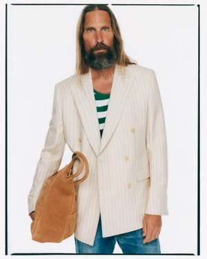 Man with long hair and beard wearing striped blazer holding a suede bag against a white background
