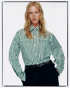 Model with long blonde hair wearing a green striped button-down shirt and black pants against a white background
