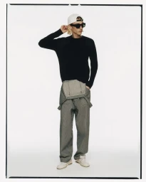 Model wearing a black sweater, striped trousers, white cap, and sunglasses against a plain studio background.