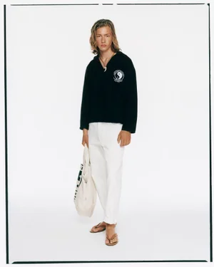Long-haired male model wearing black hoodie and white trousers holding a cream tote bag against a plain white background