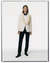 Model wearing a white tuxedo jacket, black bow tie, flower lei, and black trousers against a white seamless backdrop.