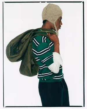 Profile of a black man wearing a green striped sweater, knitted beanie, and carrying a large canvas duffel bag against a white background