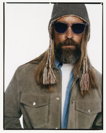 Portrait of a man with long hair, a beard, sunglasses, and a grey knit beanie against a plain white background