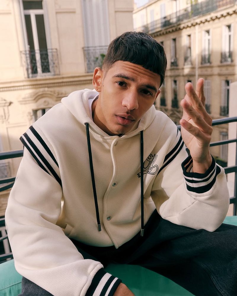 A young adult male poses on a balcony in a cream-colored hoodie with black stripes, showcasing fashion in an urban setting with natural light.