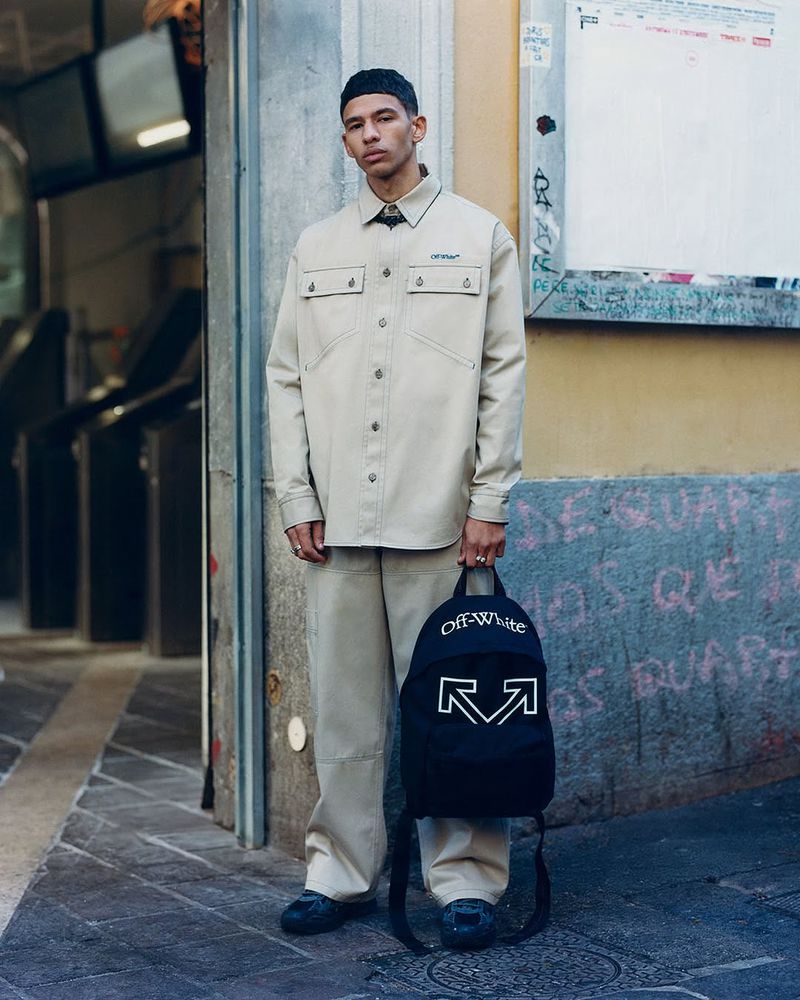 Man in neutral-toned shirt and pants poses in an urban setting while holding a black backpack with white Off-White logos.