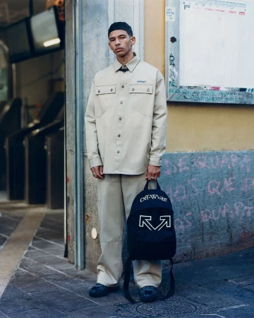 Man in neutral-toned shirt and pants poses in an urban setting while holding a black backpack with white Off-White logos.