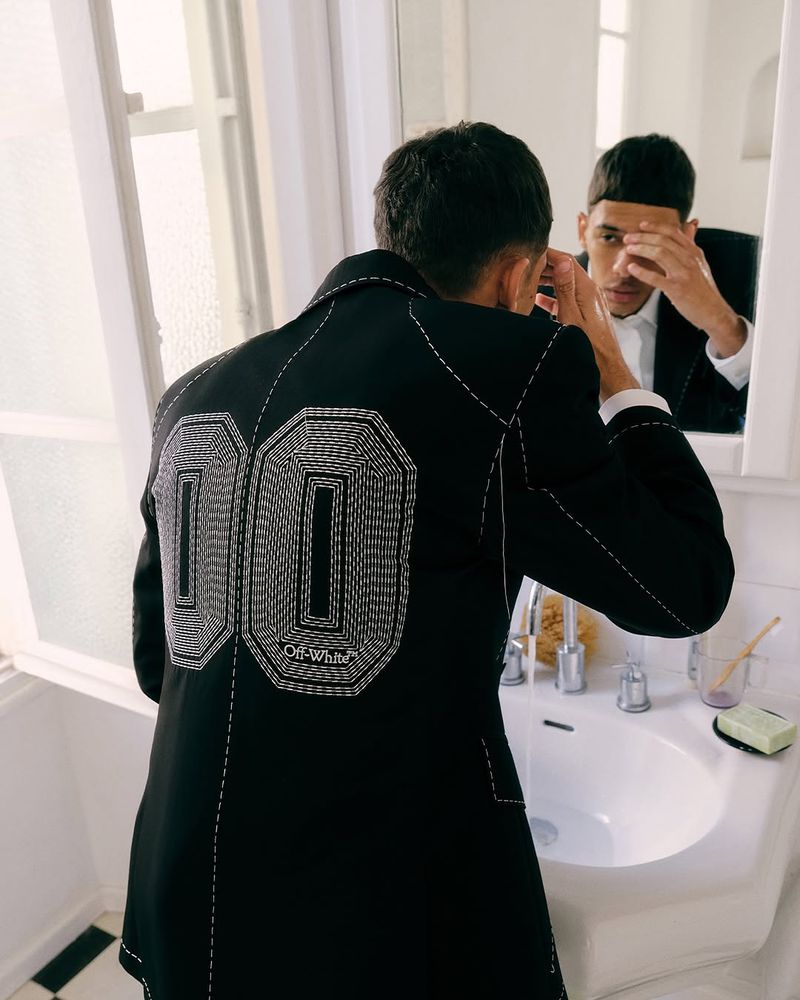 Adult male in a black jacket with white detailing, visible in the mirror while standing in a bathroom with a white sink.