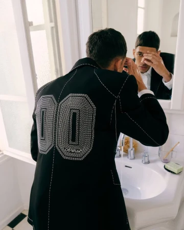 Adult male in a black jacket with white detailing, visible in the mirror while standing in a bathroom with a white sink.