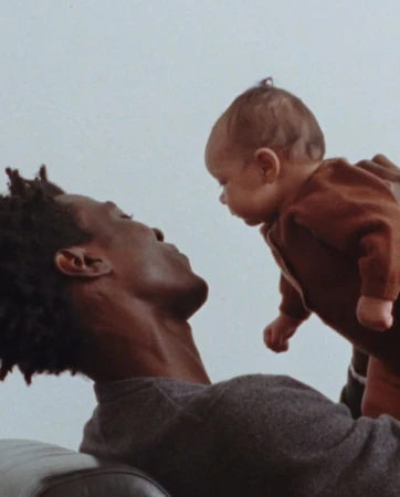 Father holding baby indoors, looking at each other. The infant wears a brown outfit. The adult male has short hair and a grey sweater.