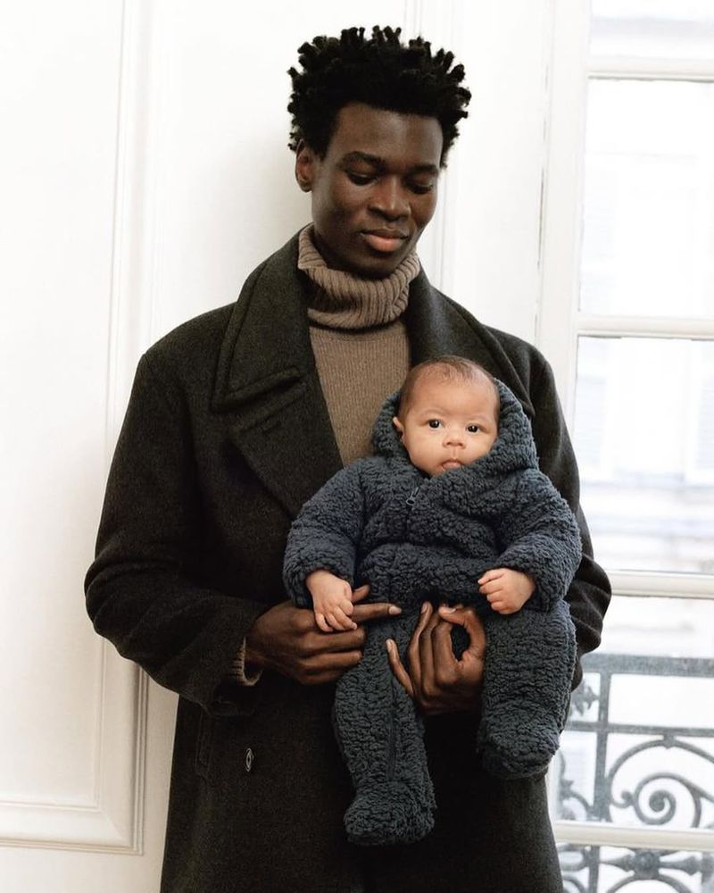 An African-American man in a coat holding a baby in a hooded snowsuit in front of a window, creating a heartwarming and stylish family moment.